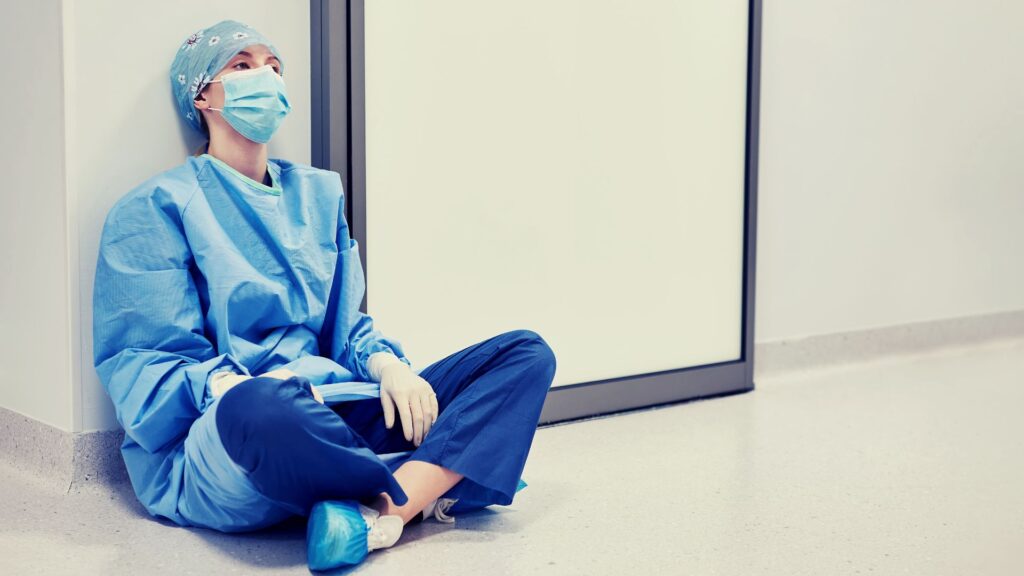 Mujer profesional de la salud, sentada en el suelo con expresión perdida y desmotivada, presentando Síndrome de Burnout.
