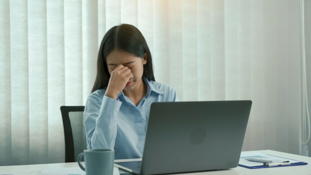 Mujer con síndrome de burnout, sentada frente a un computador con los ojos cerrados y pellizcando su nariz en gesto cansado.